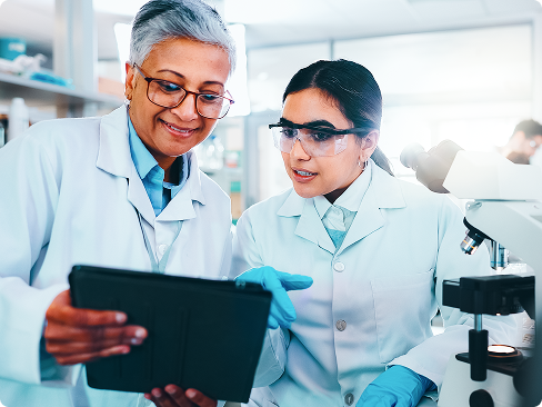 Duas pessoas de jalecos brancos trabalham em um ambiente de laboratório. Uma está segurando e olhando para um tablet eletrônico, enquanto a outra está usando luvas e está ao lado de um microscópio, aparentemente discutindo os dados ou resultados apresentados no tablet. Ao fundo, pode-se ver outro indivíduo trabalhando perto de equipamentos de laboratório.