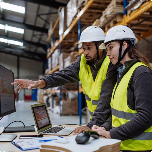 Duas pessoas em coletes de segurança refletivos e capacetes de proteção estão em pé em um armazém, trabalhando juntas em frente a um computador portátil sobre uma mesa, que também possui outros itens como papéis e um mouse. Uma pessoa está apontando para a tela do computador, possivelmente discutindo um assunto de trabalho com a colega ao lado. O ambiente ao fundo inclui estantes altas com várias caixas, indicando que eles estão em um espaço industrial ou de armazenamento.