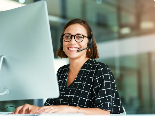 Uma pessoa sentada em uma escrivaninha em um escritório, digitando em um teclado e olhando para um monitor de computador. Ela está vestindo uma blusa de manga curta com um padrão quadriculado. A cena tem um fundo desfocado de um ambiente de escritório.