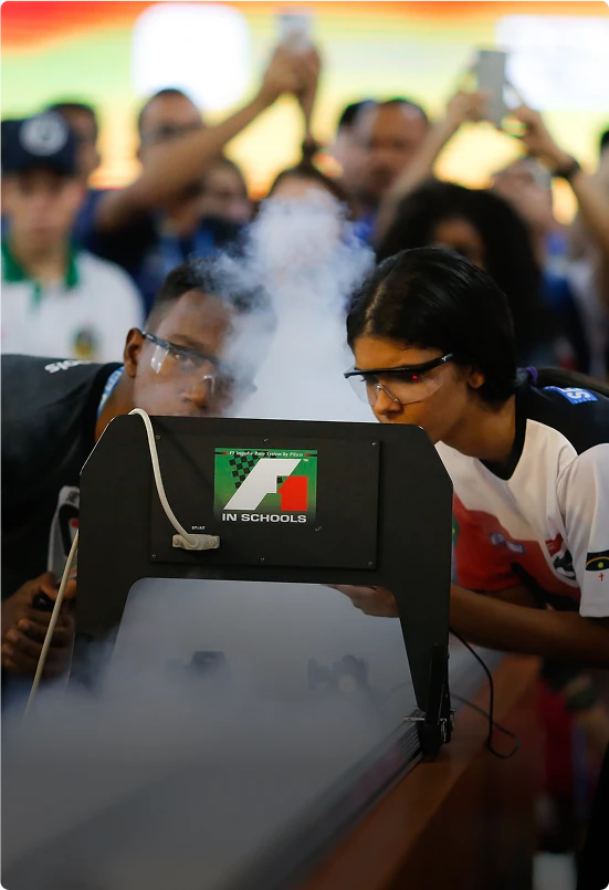 Técnico examina uma placa de circuito eletrônico com uma lupa em uma bancada de trabalho, enquanto um design de circuito é exibido no monitor do computador.